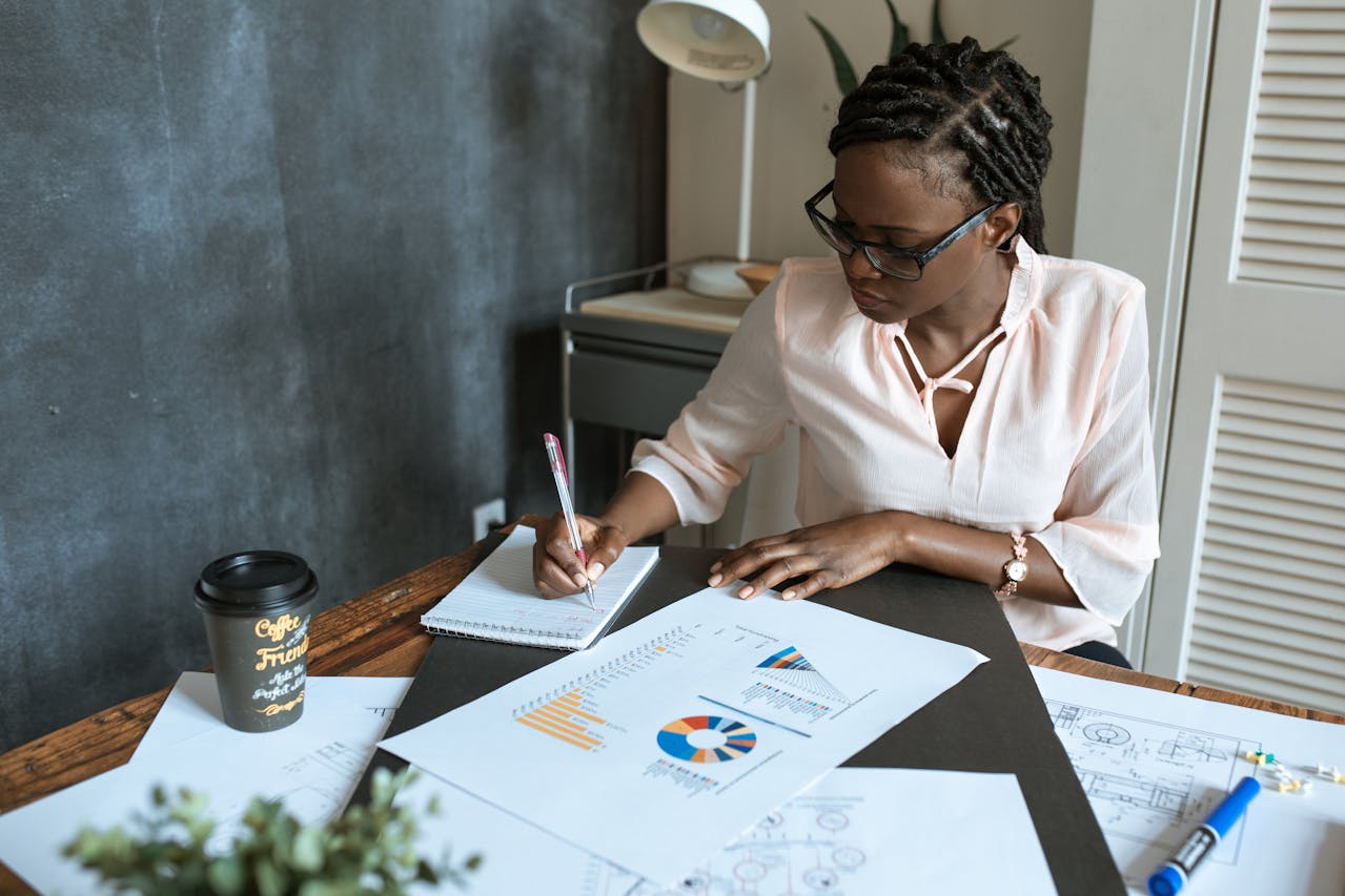 A woman is sitting at a desk with financial reports laid out in front of her and is jotting notes on one of them