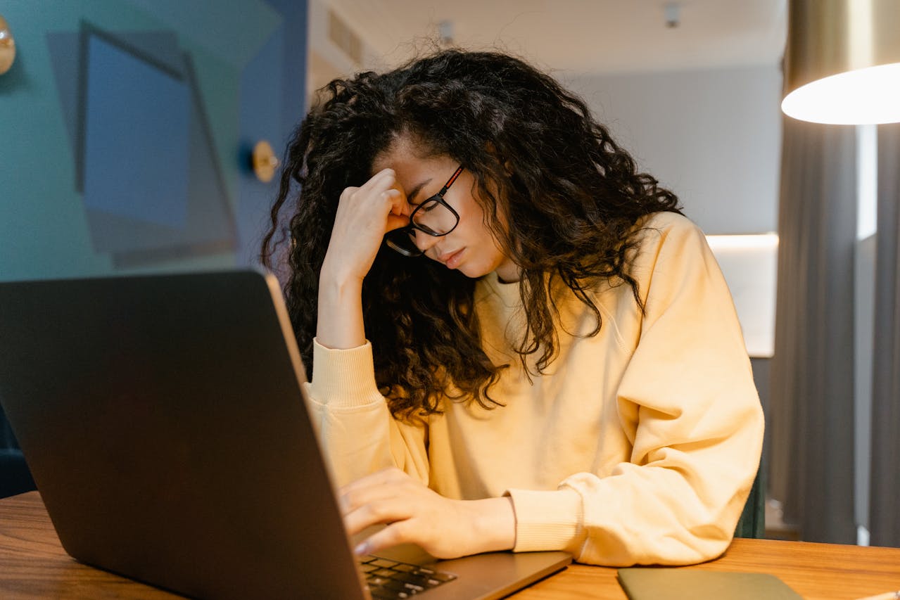A woman in a yellow sweater sits in front of a laptop with her forehead resting on her hand and an overwhelmed look on her face